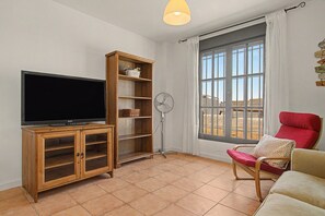 A living room with a flat-screen TV, wooden shelving, a standing fan, a window with curtains, and a red chair. - Holiday Home 'Luca' with Wi-Fi (Conil de la Frontera)