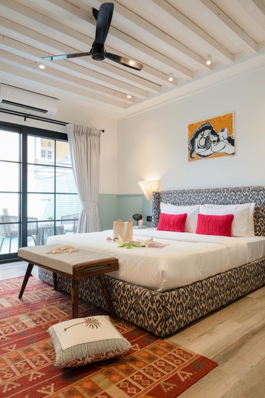 Une chambre d’hôtel avec un lit, un ventilateur de plafond, une fenêtre avec des rideaux, un tapis à motifs et une œuvre d’art accrochée au mur.