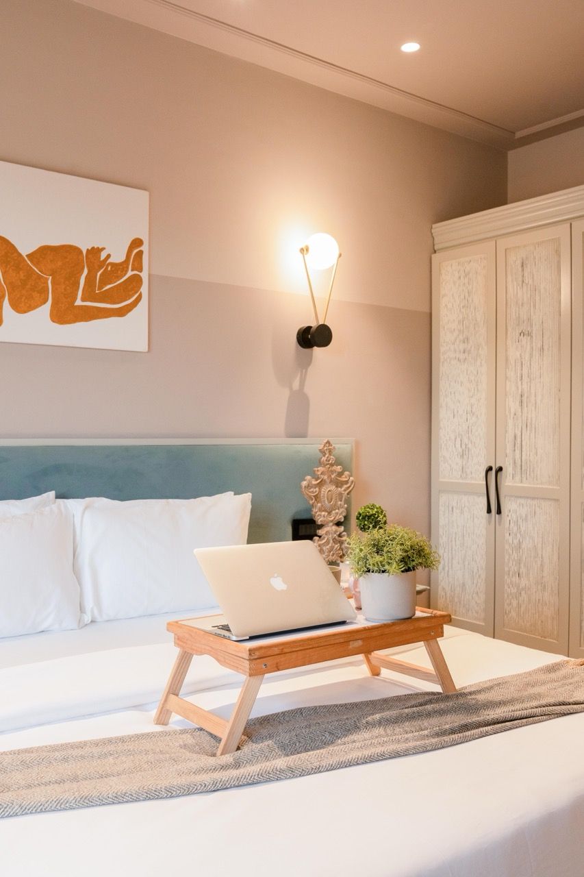 A bedroom with a bed, a laptop on a wooden stand, a wall-mounted lamp, a wardrobe, and a framed artwork.