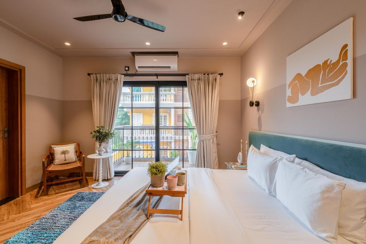 A hotel room with a bed, a ceiling fan, a window with curtains, a small table with a plant, and a wall-mounted light fixture.