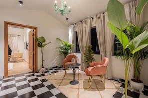 A room with a checkered floor, two pink chairs, a small round table, a chandelier, and large green plants. - Staymaster Abira-3BR Pvt Pool Villa (Assagao)