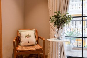 A corner room with a wooden chair, a white cushion with a palm tree design, a small white table with a vase of green plants, and a view of a balcony with a railing and buildings in the background. - Staymaster Abira-3BR Pvt Pool Villa (Assagao)