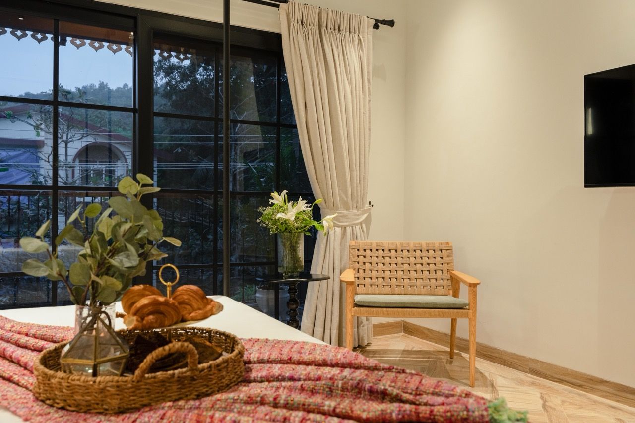 A room with a bed, a wooden chair, a window with curtains, a wall-mounted TV, and a table with a basket and flowers.