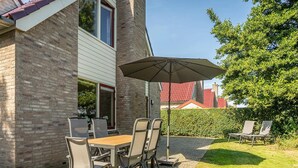 A patio with a table and chairs, a large umbrella, and a hedge. - Villa Texel With Sauna (De Koog)