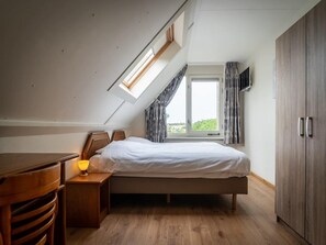 Ein Schlafzimmer mit schrägen Wänden, einem Bett, einem Nachttisch mit Lampe, einem Kleiderschrank und einem Fenster mit Vorhängen.