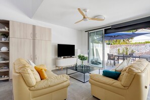 A modern living room with two beige sofas, a glass coffee table, a flat-screen TV, and a ceiling fan.