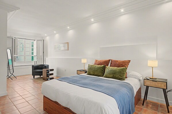 Un dormitorio con cama, mesitas de noche, una silla, una ventana y un cuadro colgado en la pared.