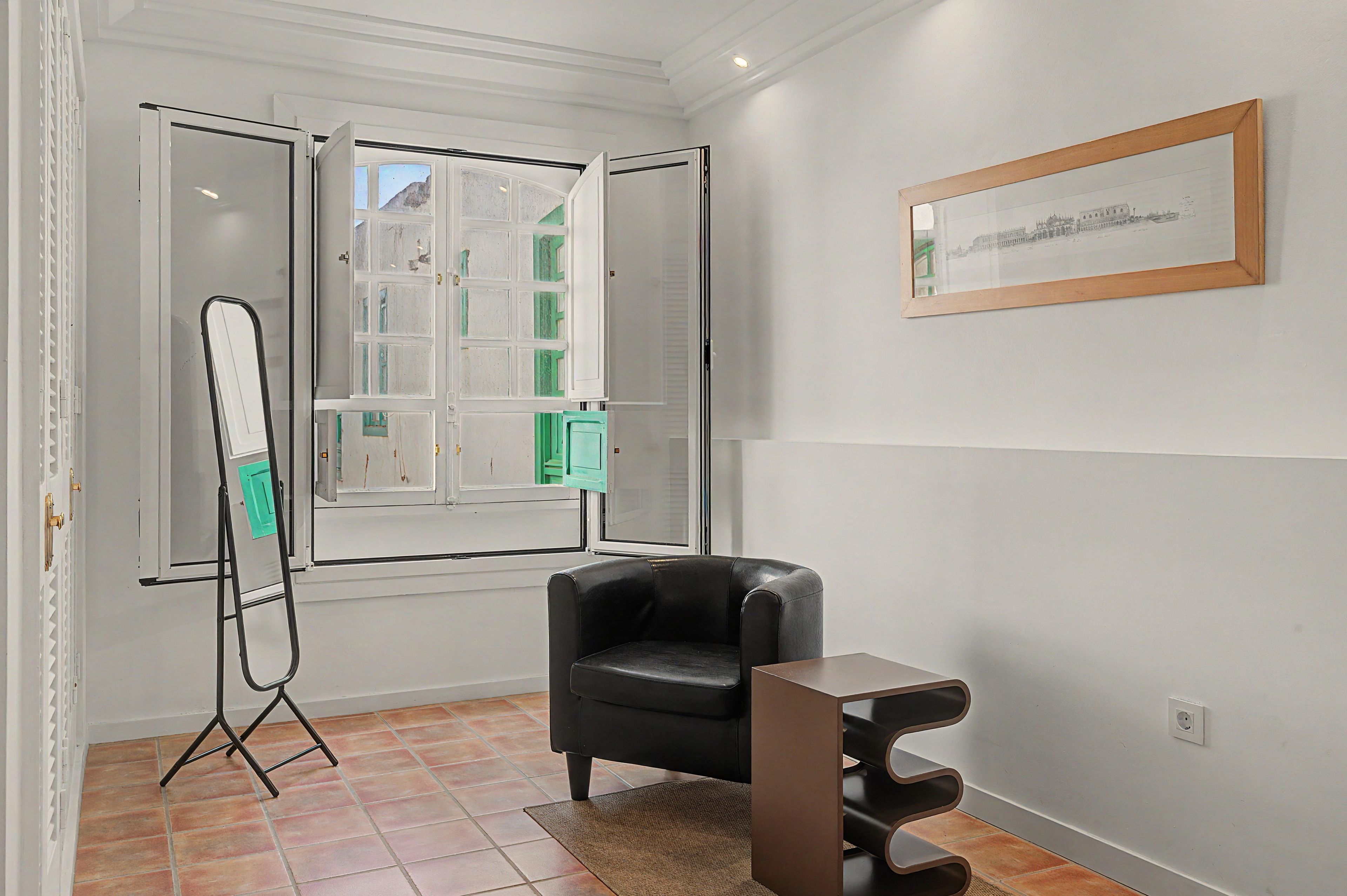 Una habitación con una butaca de cuero negro, un espejo de pie, un cuadro en la pared y una ventana con vistas a edificios.