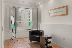 Una habitación con una butaca de cuero negro, un espejo de pie, un cuadro en la pared y una ventana con vistas a edificios.