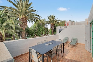 An outdoor terrace with a table and chairs, surrounded by palm trees and a building with a sign. - César’s Oasis – Manrique-Style Architecture Near Playa de las Cucharas (Costa Teguise)
