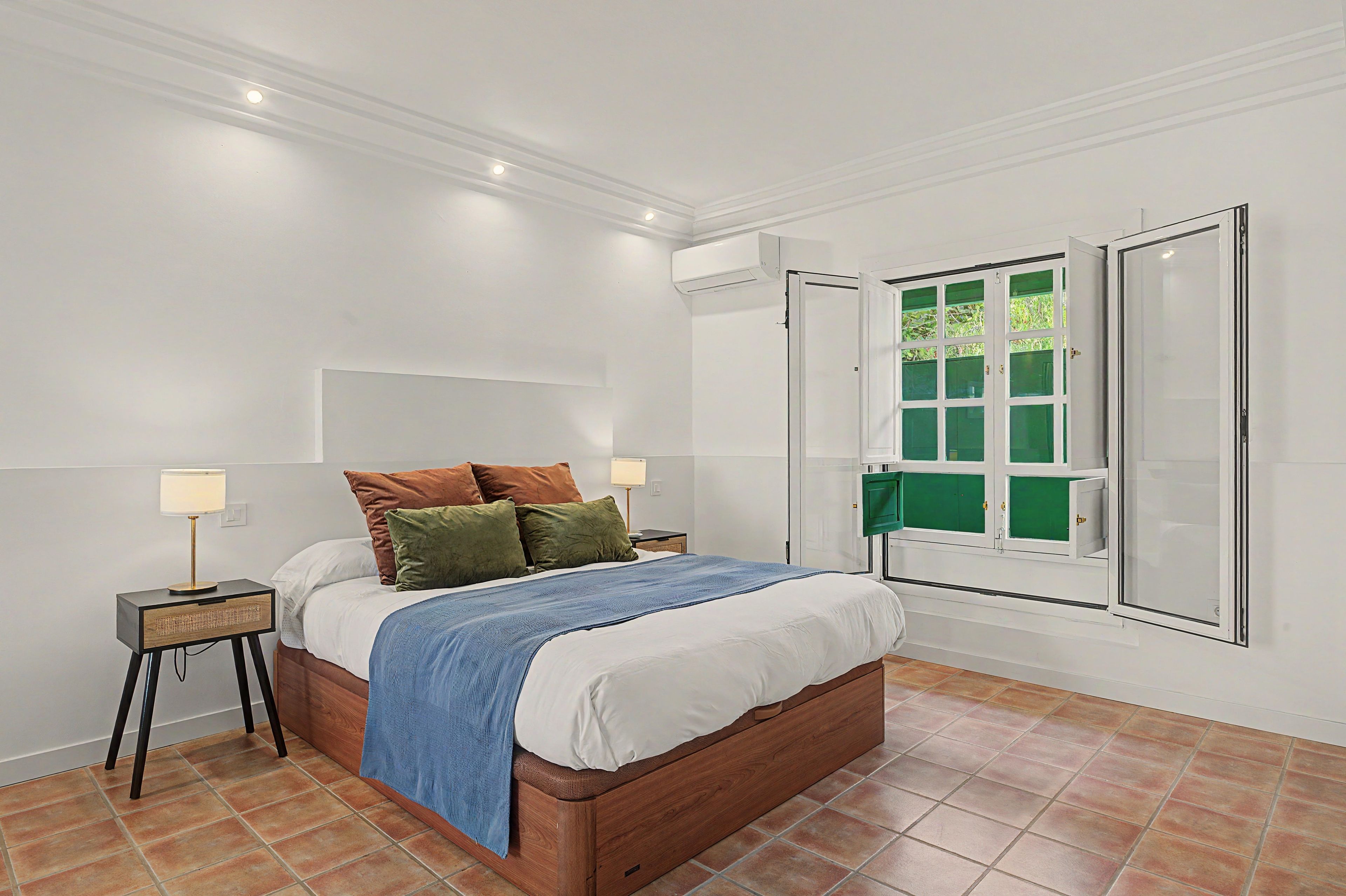 A bedroom with a bed, bedside tables, a lamp, a window with green shutters, and a white ceiling.