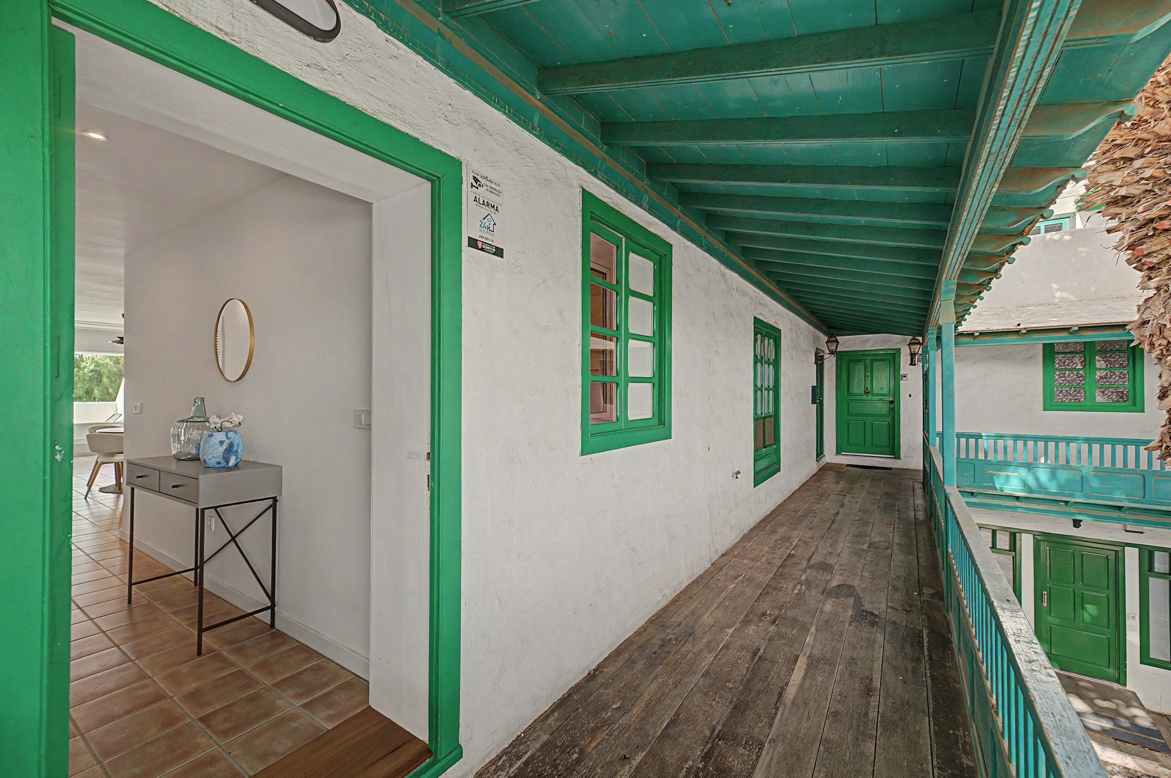 A corridor with green-framed windows and doors, a small table with a vase, and a wooden floor.