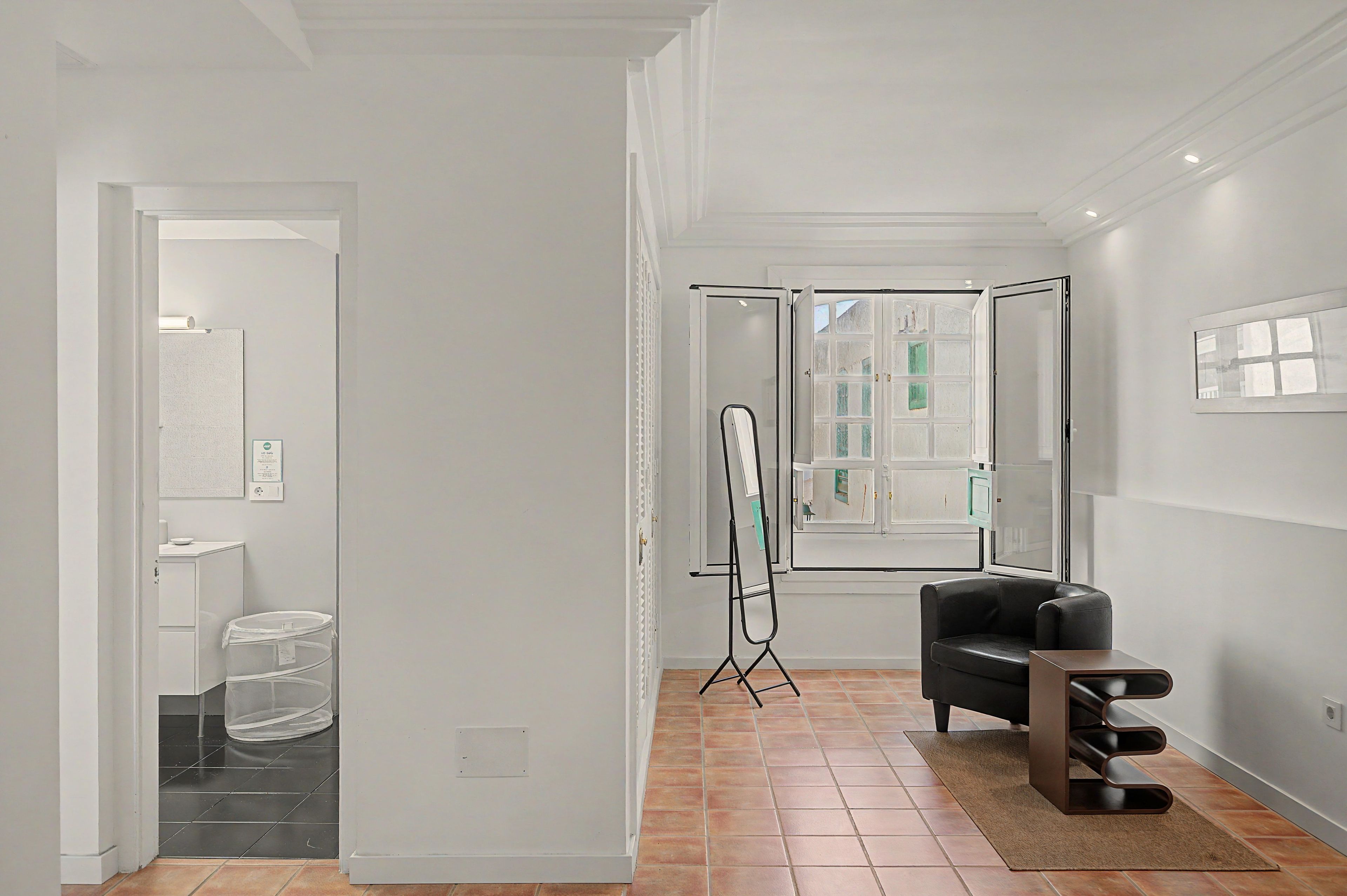 A modern interior with a black leather chair, a standing mirror, and a view of a bathroom through a glass door.