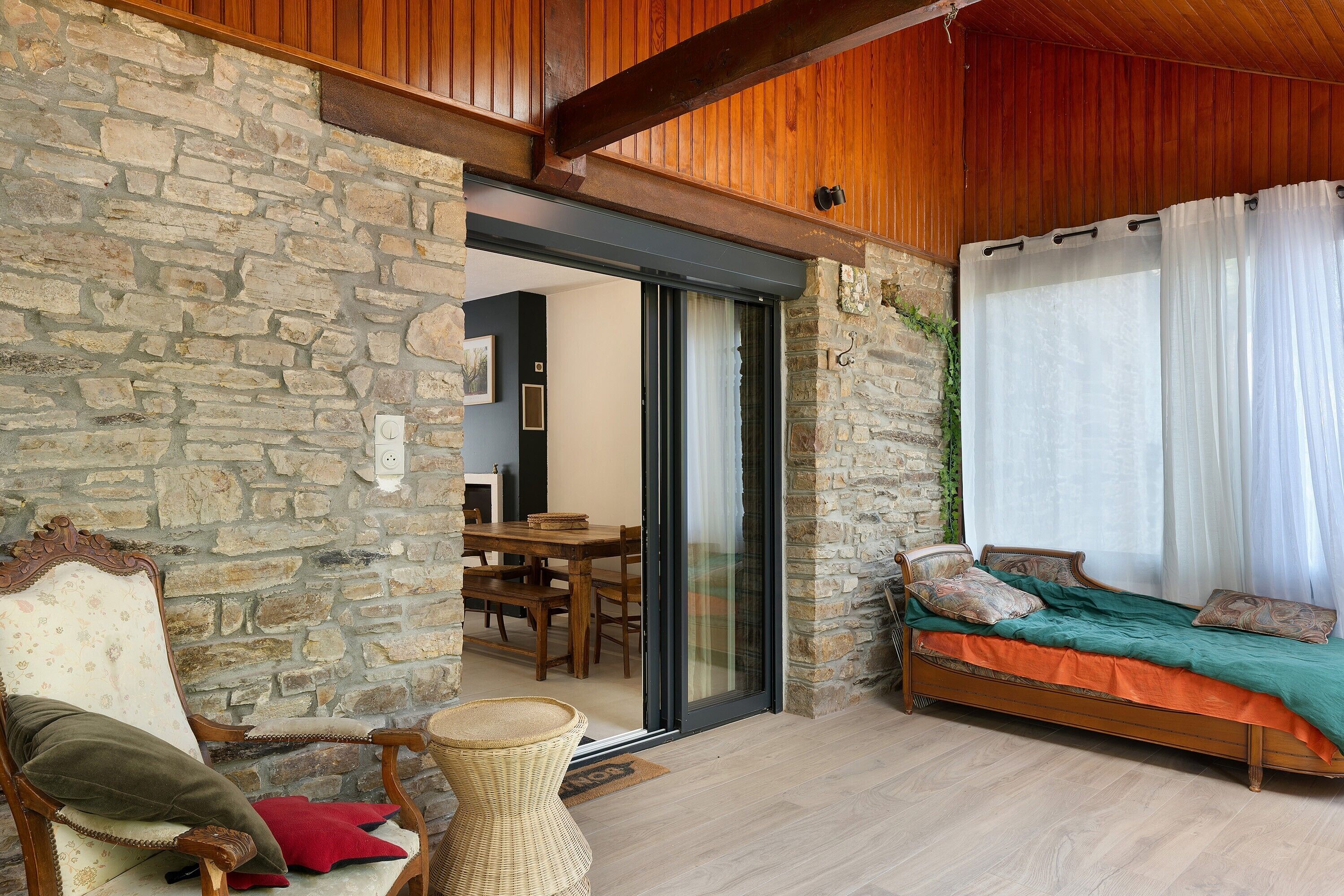 Una stanza accogliente con parete in pietra, soffitto in legno, un letto con copripiumino verde e arancione, una sedia in vimini con cuscini e un'area pranzo visibile attraverso una porta in vetro.