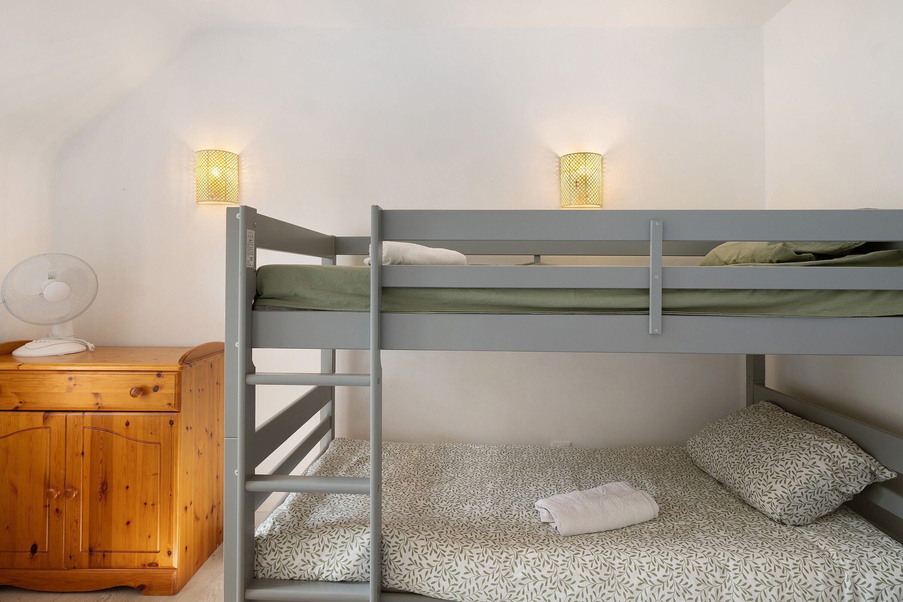 A bunk bed with two pillows and a blanket, a wooden nightstand with a fan, and two wall-mounted lamps.