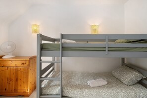 Un lit superposé avec deux oreillers et une couverture, une table de chevet en bois avec un ventilateur, et deux lampes fixées au mur.