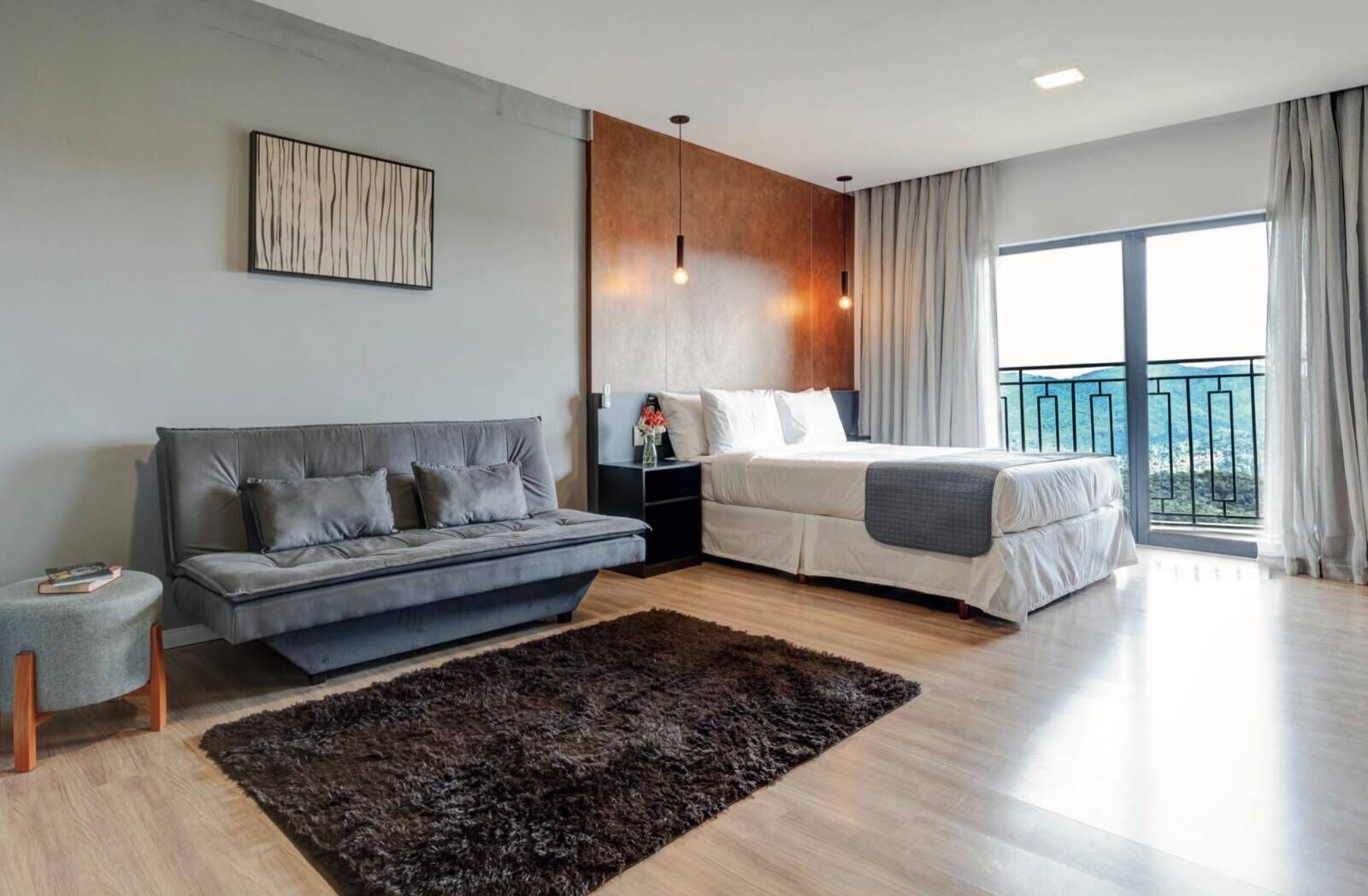 Ein modernes Schlafzimmer mit grauem Sofa, einem Bett mit weißer Bettwäsche, einem dunklen Teppich und einem großen Fenster mit Vorhängen.