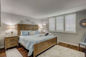 A bedroom with a wooden bed, bedside tables, a nightstand, a window with blinds, and decorative wall art. - Sunset Rentals | Island Club 6102 (Hilton Head Island)