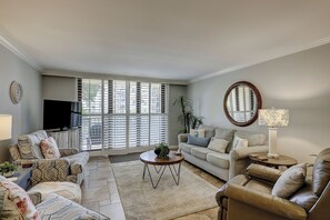 A living room with a sofa, armchairs, a coffee table, a TV, and a large window with blinds. - Sunset Rentals | Island Club 6102 (Hilton Head Island)