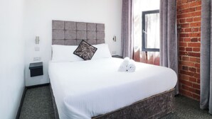 Quarto de hotel com cama, cabeceira, janela com cortinas e parede de tijolos.