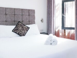 A hotel room with a tufted headboard, a floral pillow, white bedding, and rolled towels on the bed. - Apartment 304 (Nottingham)