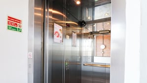 Interior de elevador com painel digital, instruções de emergência e corrimão.