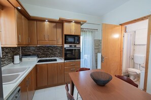 A compact kitchen with wooden cabinets, a microwave, oven, and a dining area with a table and chairs. - Venetia Beach House by Rentals Pro - Mouries Halkidiki (Mouries)