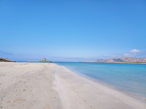 Ein Strand mit einer festungsartigen Struktur in der Ferne, klarem, blauem Wasser und einer felsigen Landschaft.