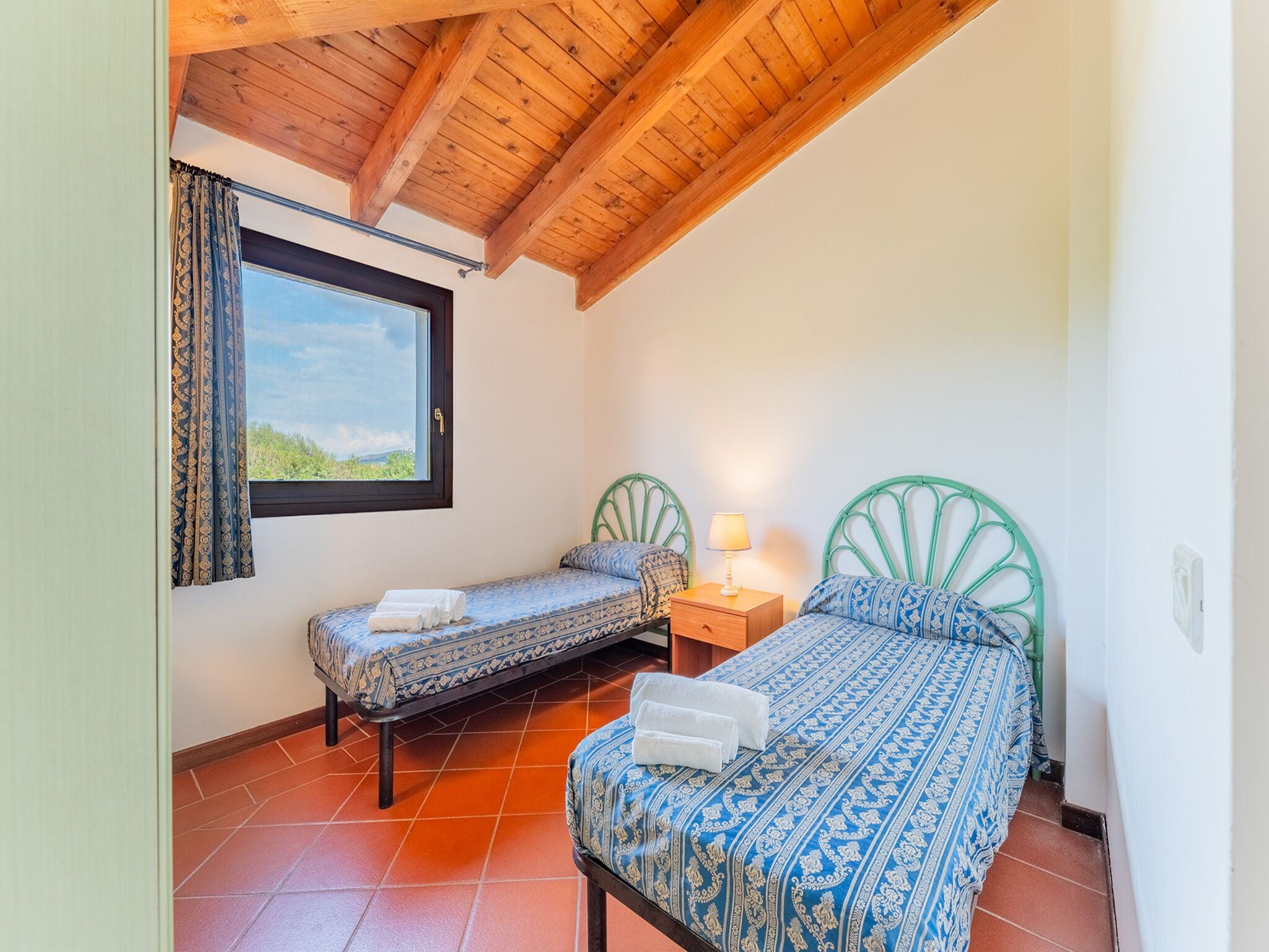 A room with two beds, a window with curtains, a wooden ceiling, and a tiled floor.