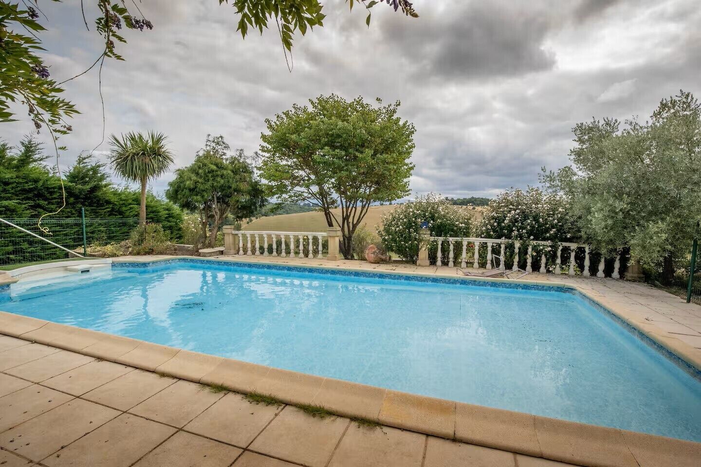 A swimming pool with a white balustrade, surrounded by trees and a cloudy sky.