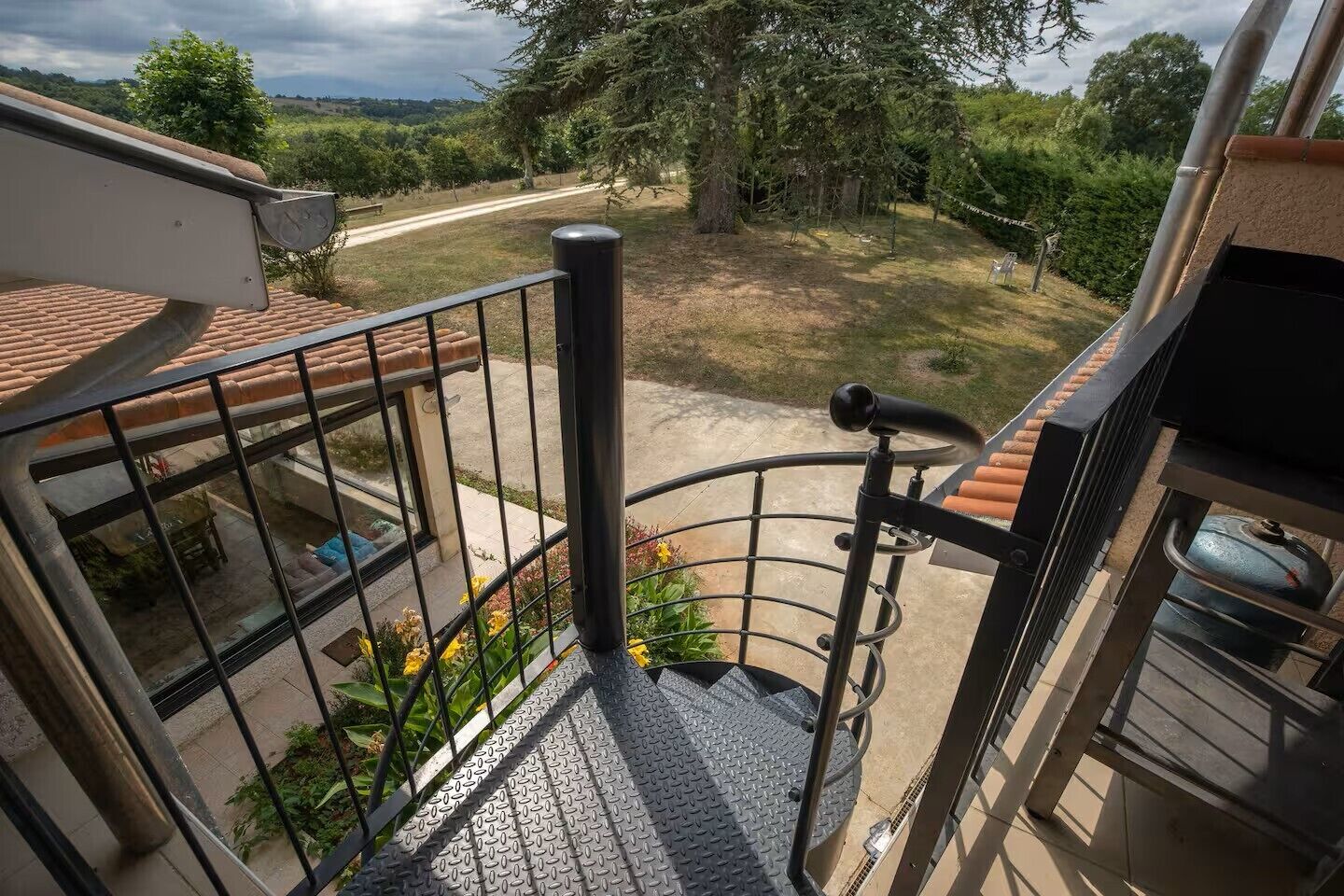 Un balcon avec une balustrade en métal, un escalier en métal et une vue sur un jardin et une route.