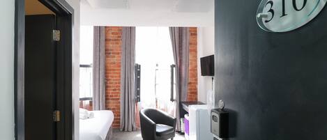 A hotel room with a bed, a black bathtub, a desk with a computer, a TV mounted on the wall, and a window with curtains.