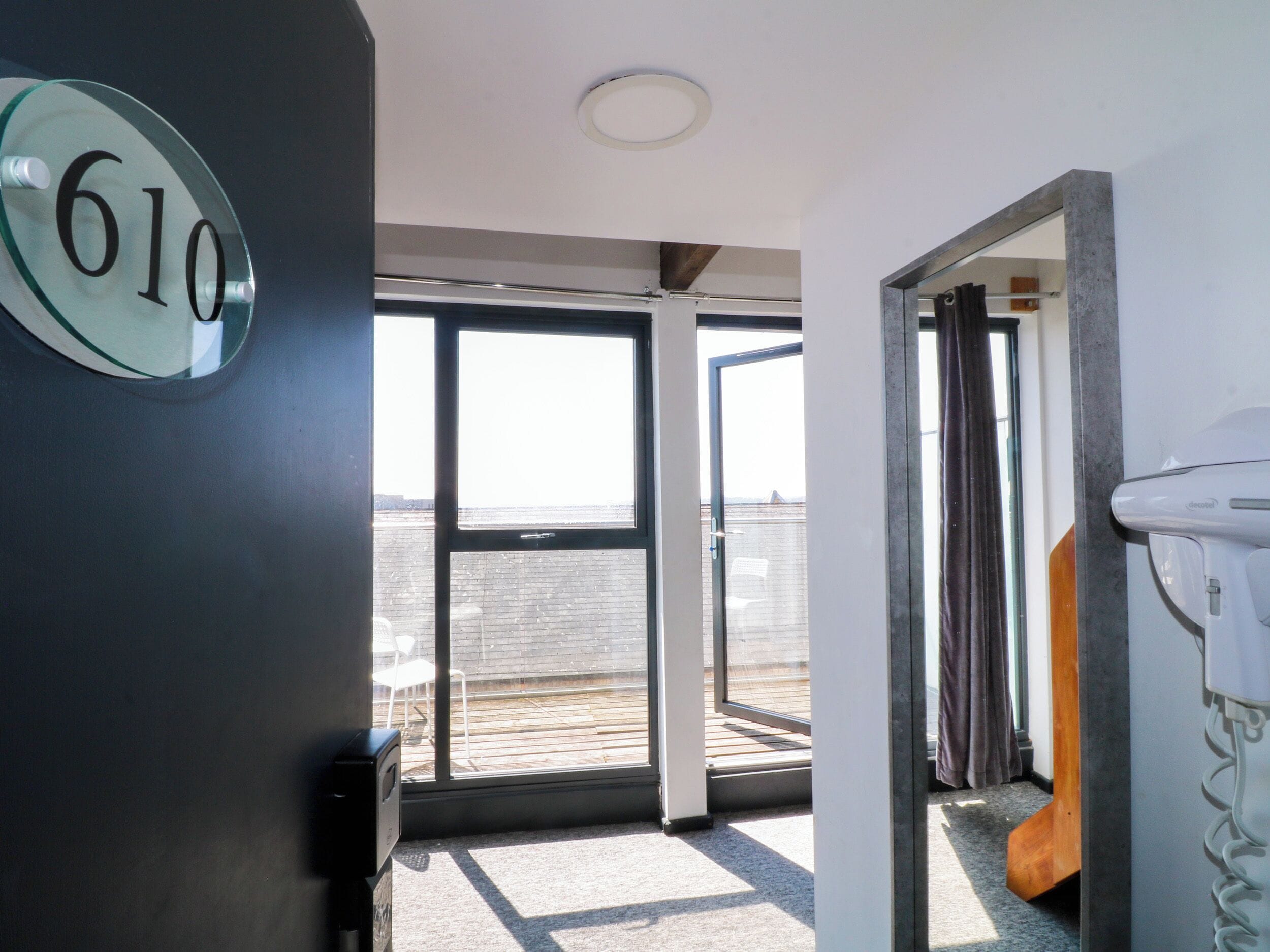 Una habitación con un amplio ventanal corredera que da a una terraza exterior, un número '610' visible en la pared y un toallero blanco.