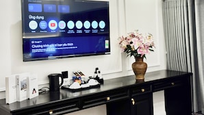 A hotel room with a flat-screen TV mounted on the wall displaying various streaming services. There is a vase with flowers on a black console table, and a tray with cups and a coffee pot. - Dollar Hotel An Khanh (Hanoi)
