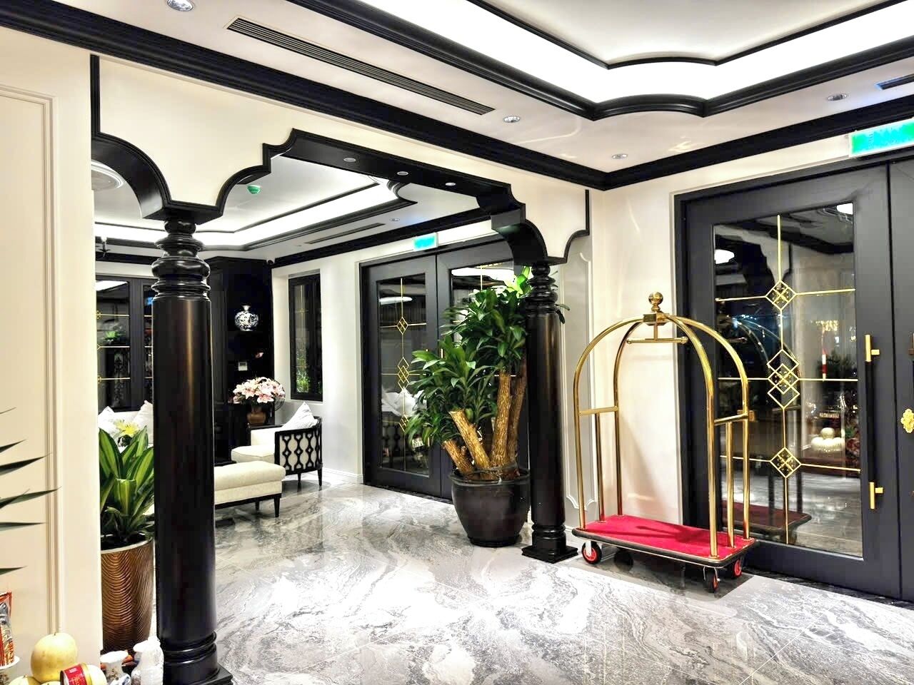 A lobby with marble flooring, a large potted plant, a gold-framed cart, and a black door with geometric patterns.