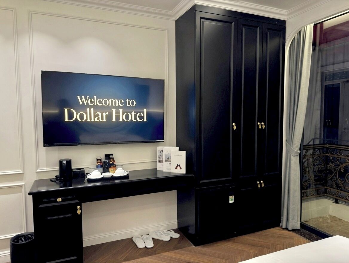 Ein Hotelzimmer mit einem großen Flachbildfernseher, der 'Welcome to Dollar Hotel' anzeigt, einem schwarzen Schreibtisch mit Kaffeemaschine, Telefon und einer Ablage mit Tasse und Buch.