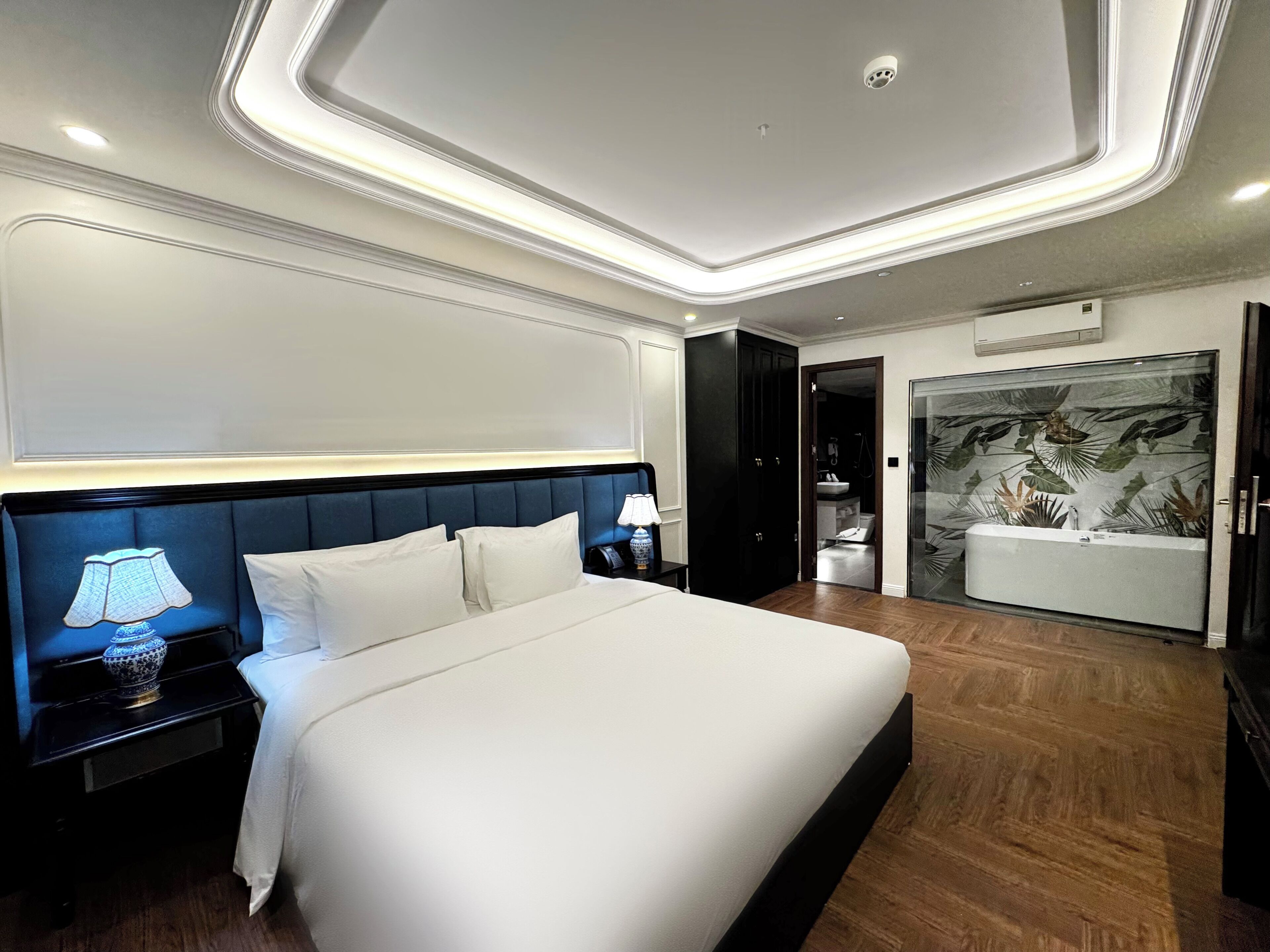 A modern hotel room with a large bed, bedside table, lamp, and a bathroom with a large bathtub.