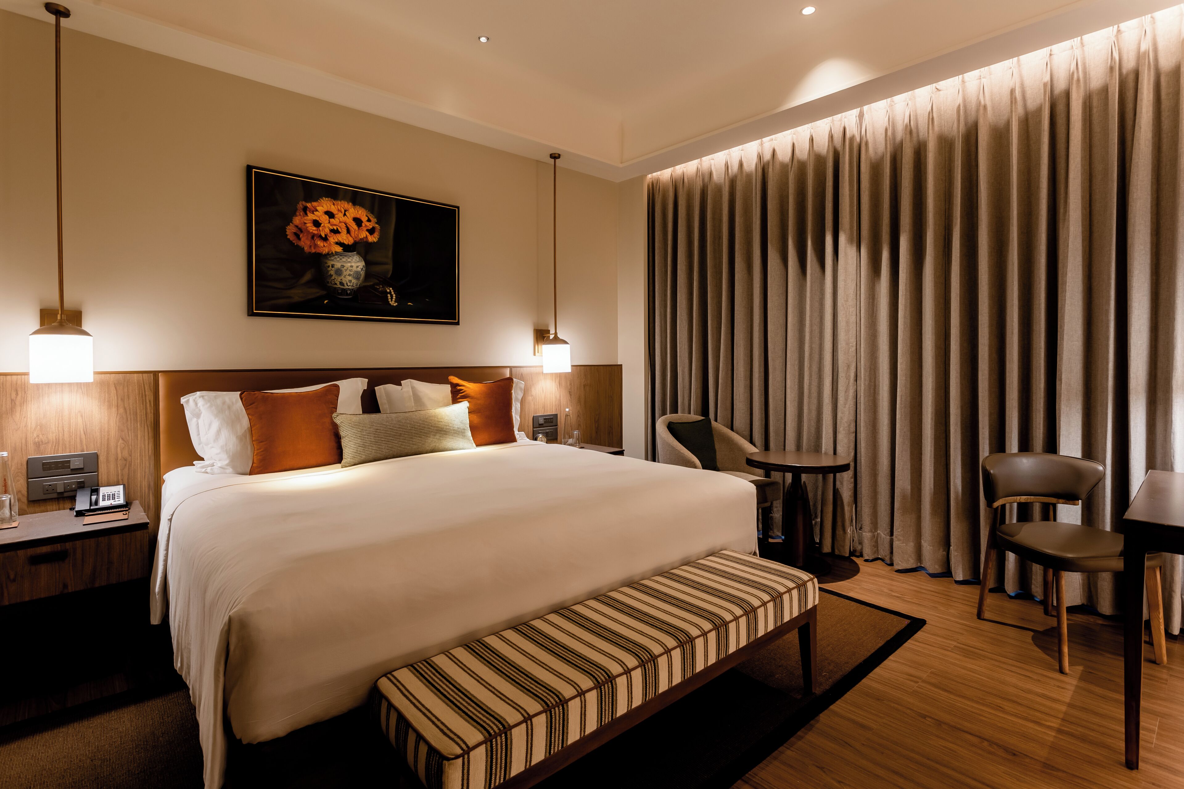Habitación de hotel con una cama grande, mesitas de noche, un escritorio, una silla, una mesita pequeña y un ventanal con cortinas.