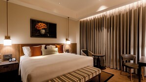A hotel room with a large bed, bedside tables, a desk, a chair, a small table, and a large window with curtains.