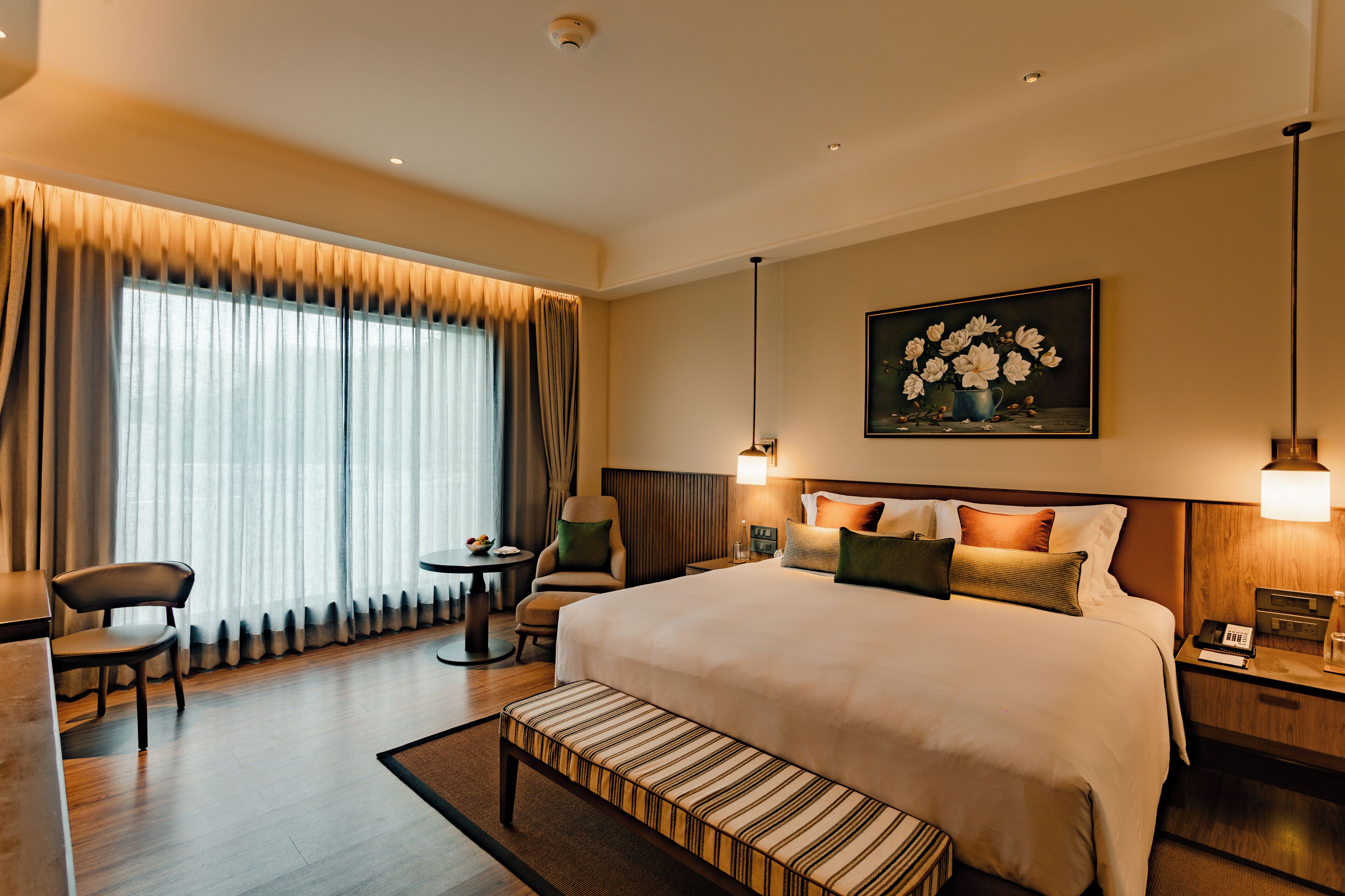 Een hotelkamer met een groot bed, een bureau, een stoel, een kleine tafel, een bankje en een schilderij aan de muur.