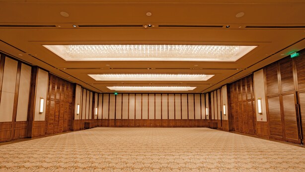 A large, empty hotel room with patterned carpet, wooden paneling, and recessed lighting.