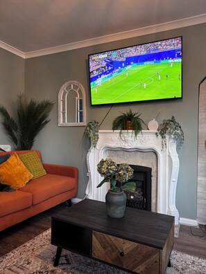 Ein Wohnzimmer mit Fernseher, der ein Fußballspiel zeigt, einem Kamin, einem orangefarbenen Sofa und einem Couchtisch.