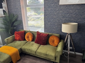 A green sofa with orange and red cushions, a floor lamp, and a large window with blinds. - Spacious Newly Reno 3BR Home with Garden | Sleeps 7 | Near BHX Airport NEC & HS2 (Birmingham)