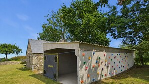 Eine Kletterwand mit bunten Griffen, die an einer Betonwand befestigt ist, ein Steinhaus und eine Rasenfläche mit Bäumen.
