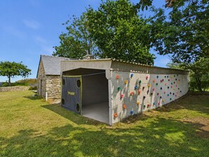 Eine Kletterwand mit bunten Griffen, die an einer Betonwand befestigt ist, ein Steinhaus und eine Rasenfläche mit Bäumen.