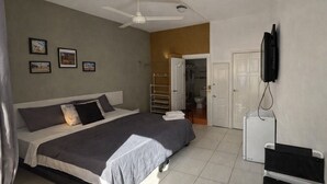 A bedroom with a bed, a ceiling fan, a TV mounted on the wall, and a door leading to a bathroom. - Room-balcony Street View Bedroom Zona Co (Santo Domingo)