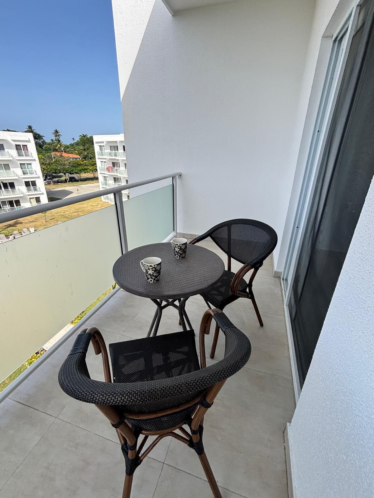 A balcony with a round table and two wicker chairs, a glass railing, and a view of buildings in the background.