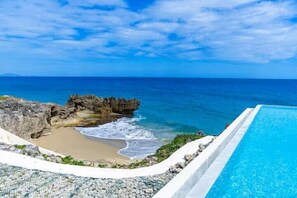 Ein Pool neben einem felsigen Strand mit Wellen, unter einem klaren Himmel.