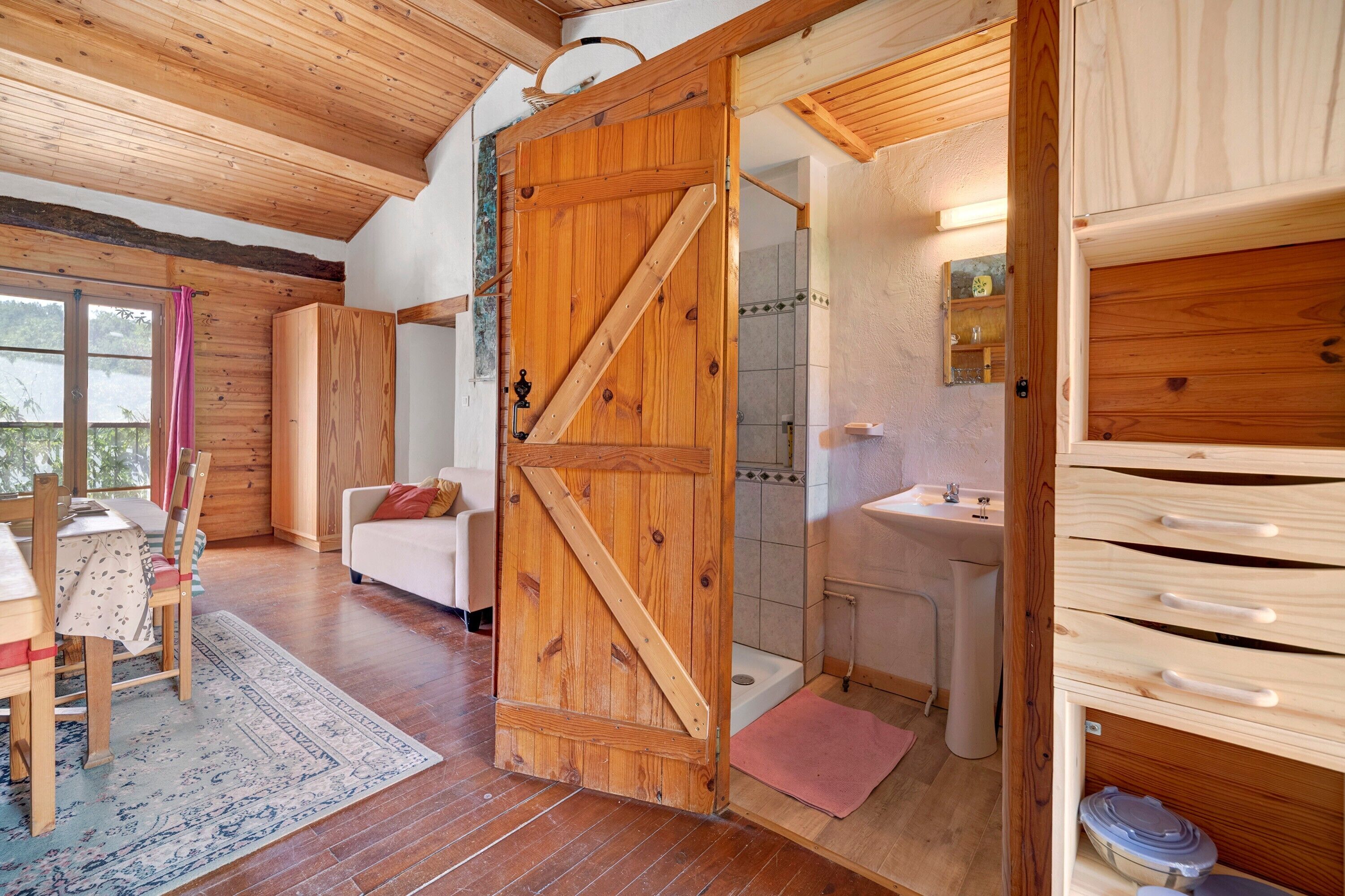 Un intérieur en bois avec une porte coulissante donnant sur une salle de bain, un coin repas avec une table et des chaises, et une vue par une fenêtre.