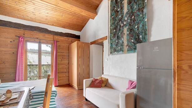 Une pièce chaleureuse aménagée comme une cabane, avec un plafond en bois, une grande fenêtre avec des rideaux, un canapé blanc, un meuble en bois et un réfrigérateur.